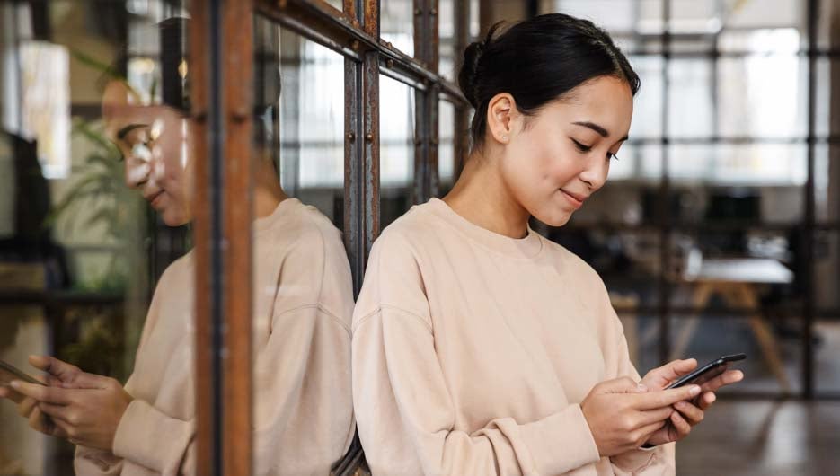 Image of young beautiful asian woman smiling and holding cellphone while working in office