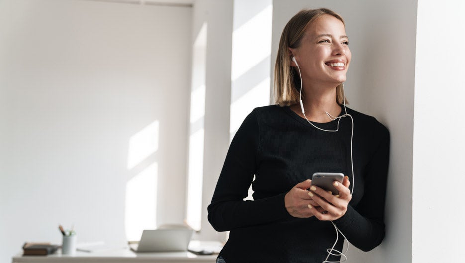Image of a beautiful optimistic young blonde business woman in office indoors listening music with earphones using mobile phone.