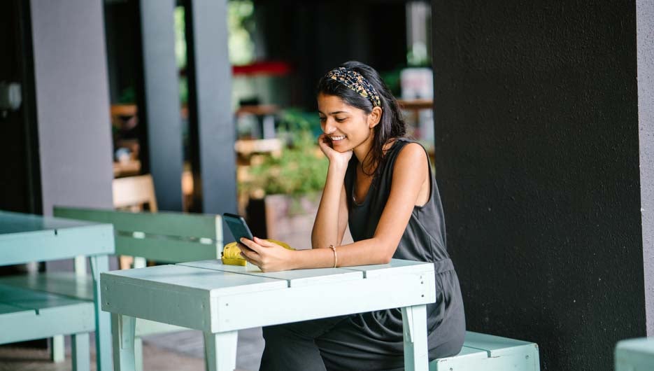 portrait of young woman sitting and resting her arms on a table at a cafe.  She appears to be looking at her smartphone.