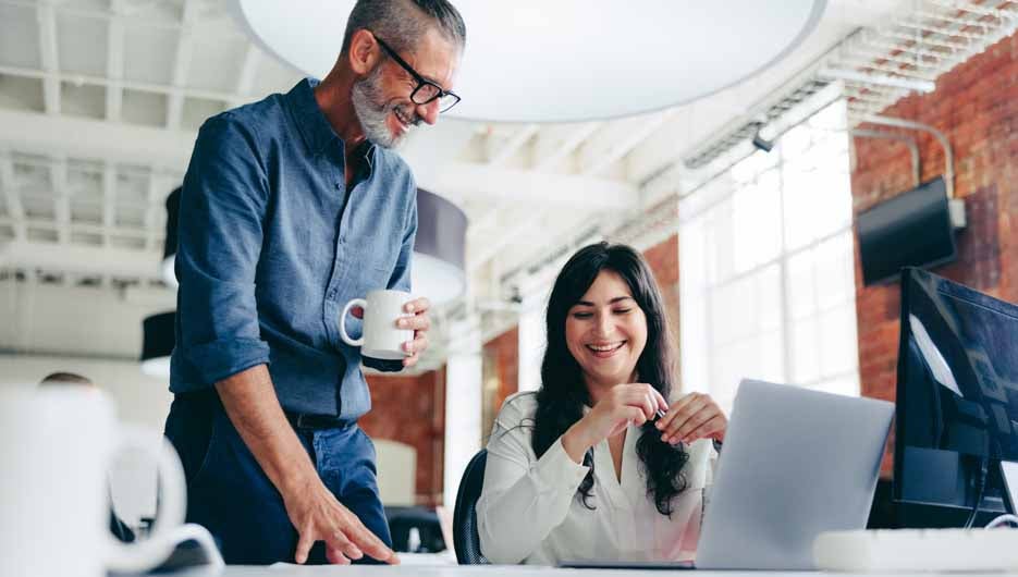 Two happy colleagues collaborating on a new project in the office. Mature businessman having a discussion with his female colleague while standing at her desk in a modern workplace.