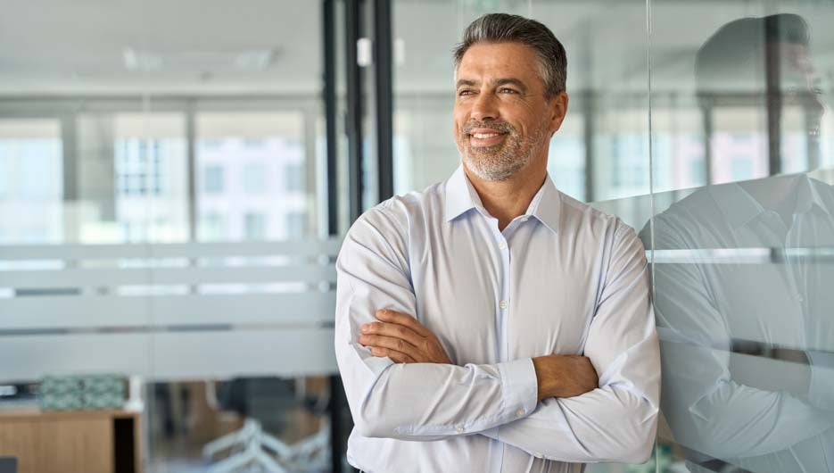 Happy mid aged older professional business man, mature latin executive manager, 50 years old investor standing in office arms crossed leaning at glass wall looking away thinking of success concept.
